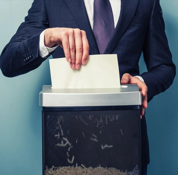 A person in a suit feeds a sheet of paper into a commercial paper shredder.